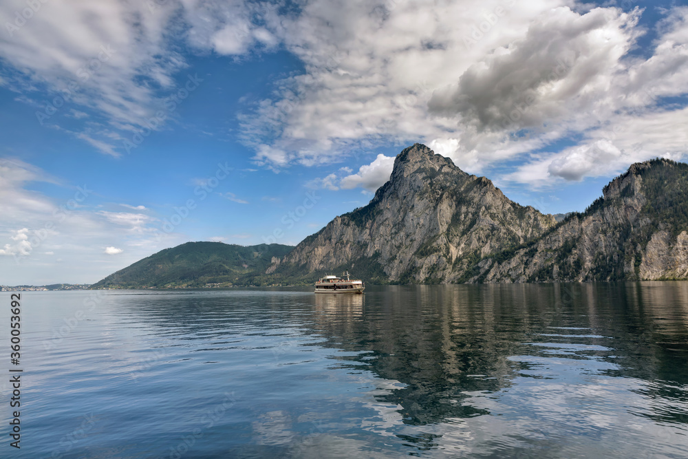 View from Traunkirchen over Traunsee lake to excursion boat and Traunstein mountain, reflection of mountains and cloudy sky in the water. Salzkammergut, Upper Austria, Europe
