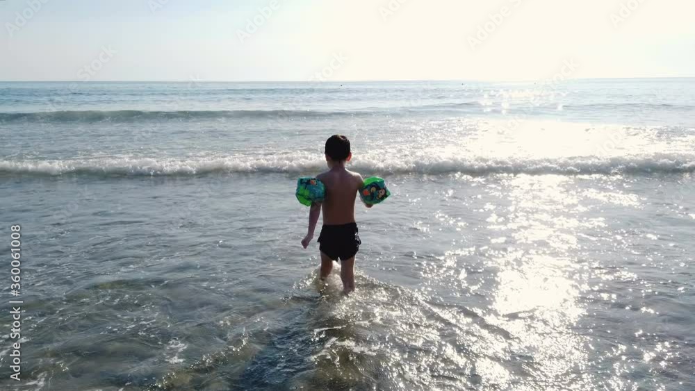 Happy child son with water wing walking to sea horizon over splashing waves,summer vacation lifestyle Happy child son with water wing walking to sea horizon over splashing waves,summer vacation lifestyle