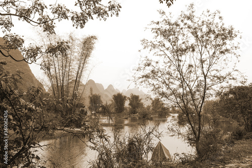 Pastoral scene of lacy trees lining Li River in China with karst mountains in the background. 