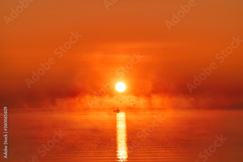 Foggy red sunrise over water reservoir in Zatoka Pucka bay located in Puck, Poland. Lonely fisherman on the boat fishing at dusk in summer.  photo