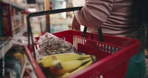 Close up of female customer carrying on red basket full of high quality products at grocery store. Happy woman doing regular shopping of goods at local market