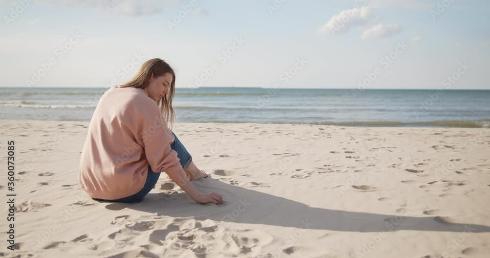 Young hipster woman sitting on a beach stroking sand by hand Young hipster woman sitting on a beach stroking sand by hand