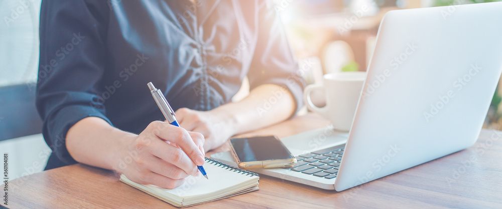 Woman is writing on a notebook with a pen and using a laptop to work in the office.