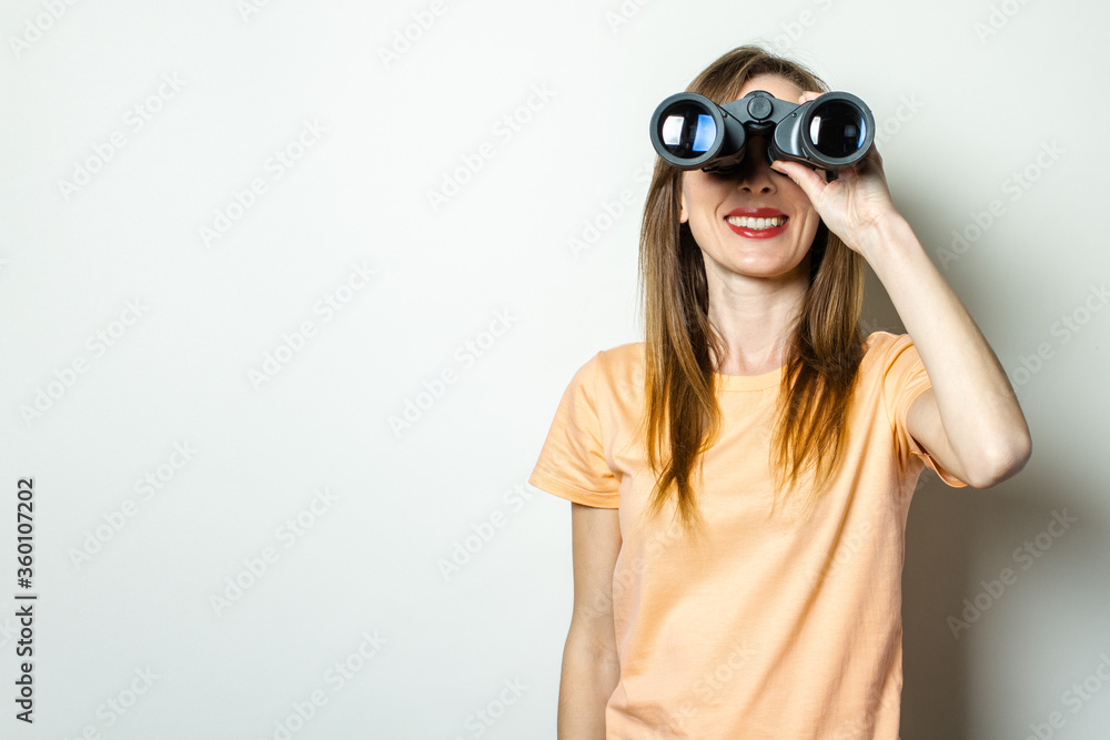 Young friendly girl looking through binoculars on a light background. Banner. Emotional face Young friendly girl looking through binoculars on a light background. Banner. Emotional face