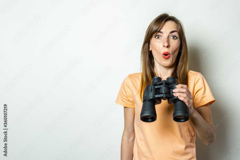 Young friendly girl with a surprised face in a T-shirt is holding binoculars on a light background. Banner. Emotional face Young friendly girl with a surprised face in a T-shirt is holding binoculars on a light background. Banner. Emotional face