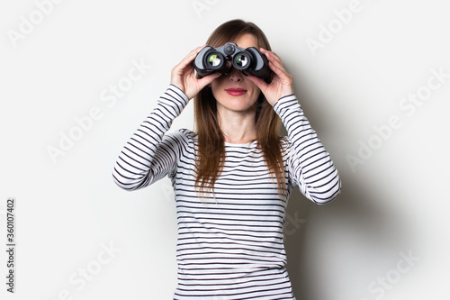 Young friendly girl looking through binoculars with the back side on a light background. Banner. Emotional face