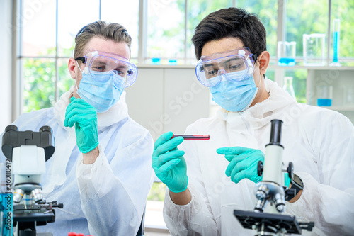 Couple male scientist wearing protection suit holding vaccine and working with many lab equipment for research vaccine at laboratory.