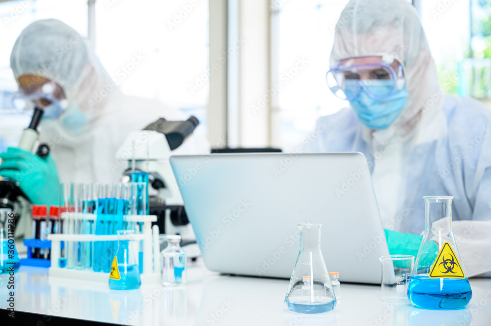 Lab equipment with blue liquid inside stand on the table with couple male scientist wearing protection suit working with computer laptop for research at laboratory at the background. Lab equipment with blue liquid inside stand on the table with couple male scientist wearing protection suit working with computer laptop for research at laboratory at the background.