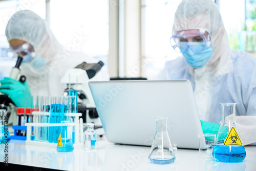 Lab equipment with blue liquid inside stand on the table with couple male scientist wearing protection suit working with computer laptop for research at laboratory at the background.