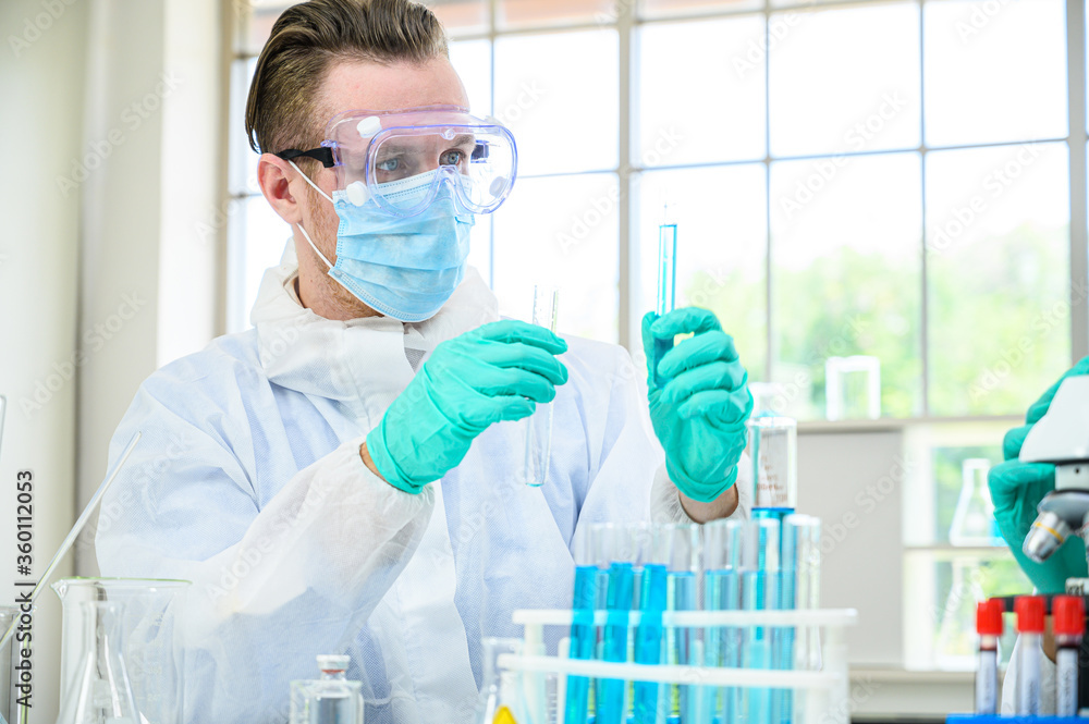Male scientist wearing protection suit holding beaker or tube with sample liquid inside and working with many lab equipment for research at laboratory. Male scientist wearing protection suit holding beaker or tube with sample liquid inside and working with many lab equipment for research at laboratory.