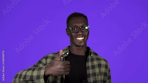 Happy, smiling african american man in good mood on blue background. Man in stylish glesses gesturing with thumbs up, like gesture and tooty smiling - human emotions photo