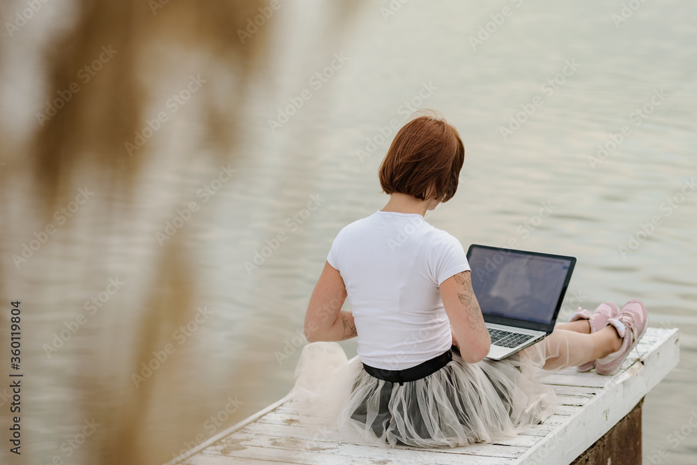 Back view of young woman sitting on pier and using laptop