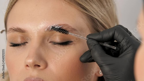 Master applies brow paste with a brush to eyebrows. Beautiful attractive female face of a blonde well-groomed woman or lady. Styling and lamination of eyebrows. Stylist's hands in black gloves photo