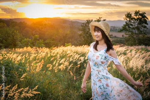 The girl wearing a hat, wearing a white dress, stand in the middle of the grass with beautiful white flowers with a relaxed and happy mood on mountain in sun set time.