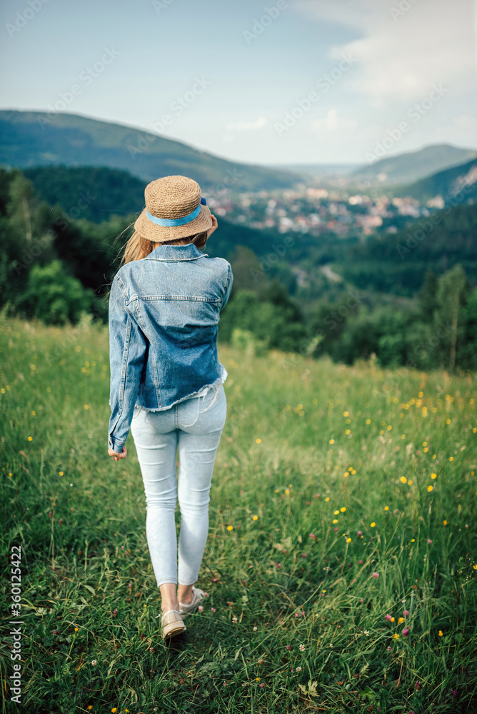 A young girl stands on a meadow on top of a mountain and looks at the mountains, holding a straw hat. Vertical photo of a girl on a background of mountains