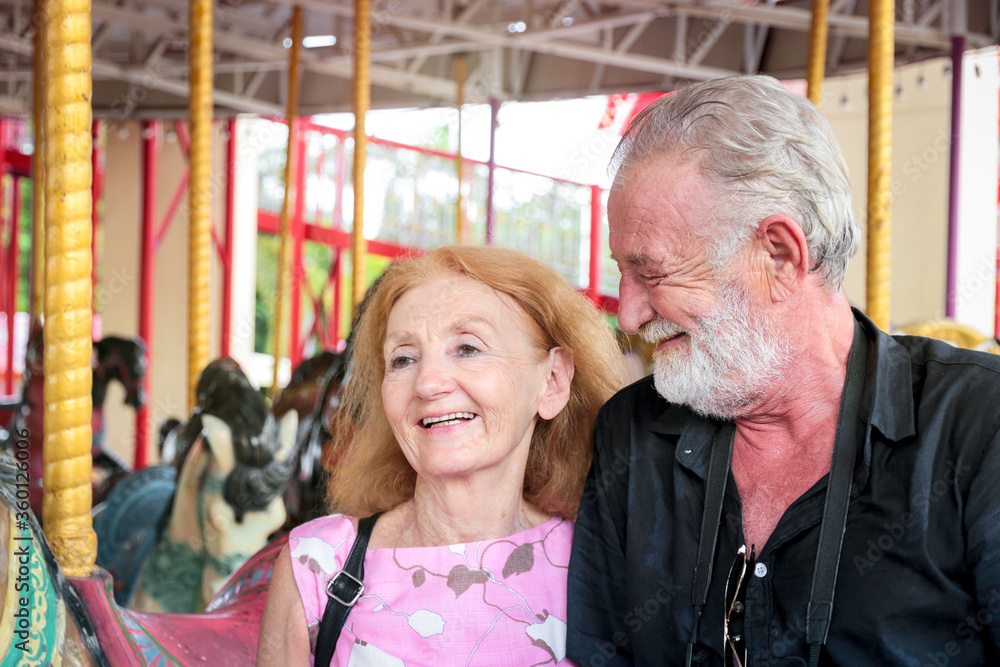 Obraz premium Elderly couple spending time together at theme park on weekend, senior people hanging out and having fun at amusement park. Grandpa and grandma enjoying at carousel spinning