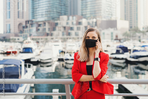 Photography Young woman in red suit wearing reusable face mask of black color standing in city center and making a call via a smartphone