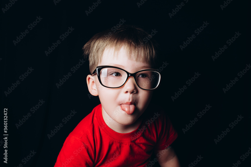 Pretty blonde boy sitting on black background in glasses, Myopia. Kid's vision, Showing tongue Pretty blonde boy sitting on black background in glasses, Myopia. Kid's vision, Showing tongue