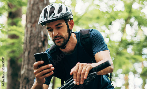 Fototapeta Naklejka Na Ścianę i Meble - Professional cyclist resting in the forest, holding mobile phone, using online application for searching GPS coordinates while riding the bike. Travel, sport and modern technology concept.