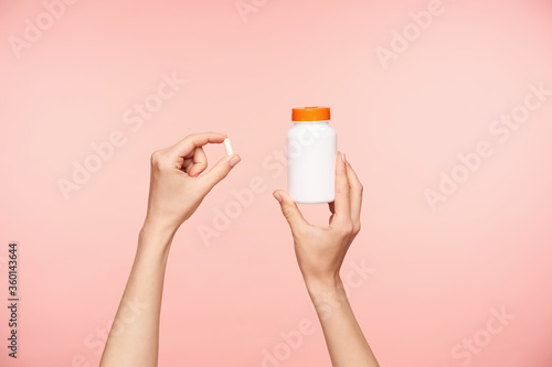 Cropped photo of raised female's well-groomed hands holding white pill and bottle with orange cover, taking vitamins while posing over pink background