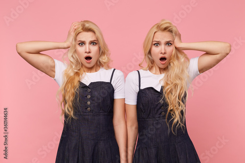 Shocked young lovely long haired blonde women keeping raised palms on their heads while looking dazedly at camera with wide mouths opened, isolated over pink background
