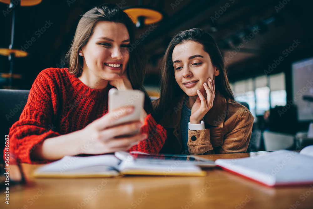 Positive female friends searching information for education using smartphone device and wifi connection in cafeteria, successful students enjoying break of studying watching funny video on telephone Positive female friends searching information for education using smartphone device and wifi connection in cafeteria, successful students enjoying break of studying watching funny video on telephone