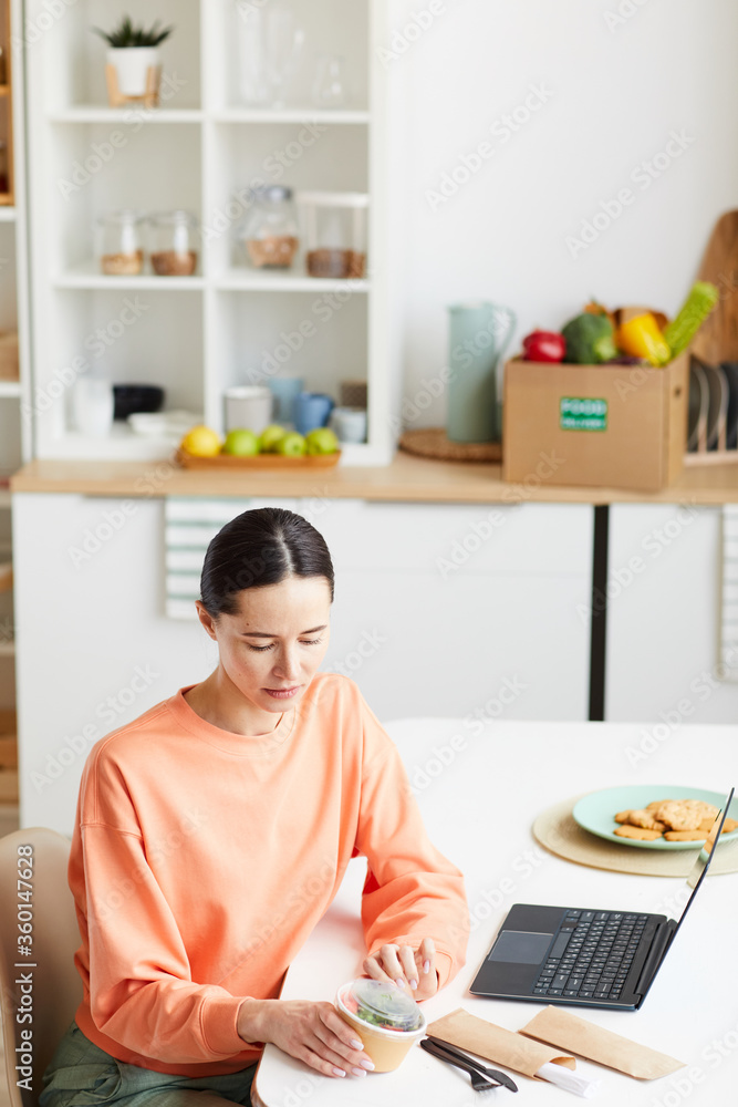 Young woman sitting at the table in front of laptop and opening the box with food in the kitchen at home Young woman sitting at the table in front of laptop and opening the box with food in the kitchen at home