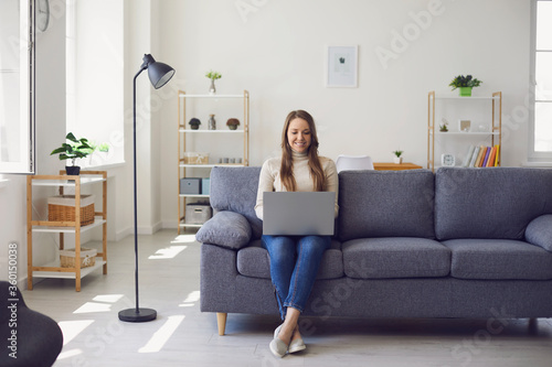 Young beautiful girl uses a laptop while sitting on a sofa in the apartment. Woman works office at home.