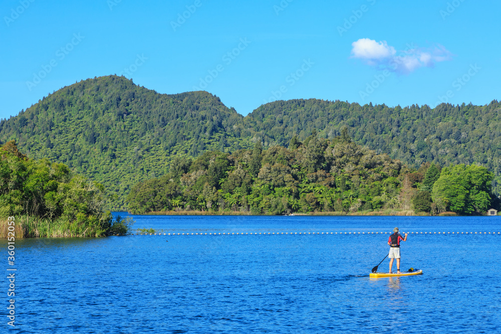 Lake surrounded by forest with a paddle boarding figure on the water. Photographed at Lake Okareka near Rotorua, New Zealand