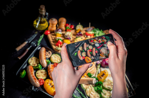 Hands taking photo a grilled sausage and mixed vegetables on a dark background with smartphone