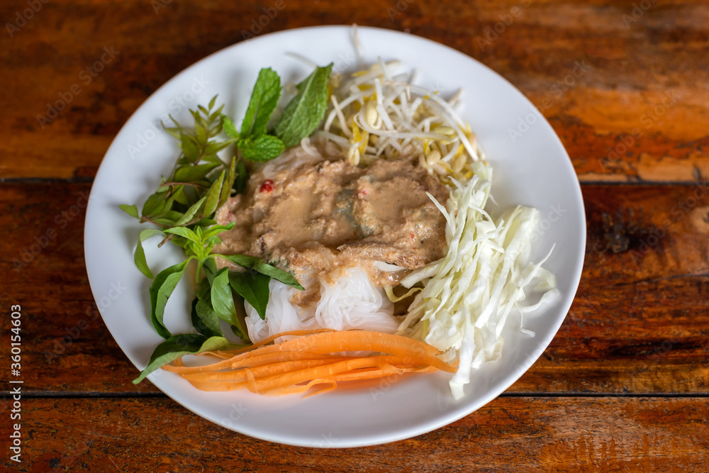 Rice Noodle eaten with curry and fresh vegetable in a white plate on a wooden table background. Fermented Rice Flour Noodles/kanom jeen are fresh rice noodles in Thai cuisine which are made from rice. Rice Noodle eaten with curry and fresh vegetable in a white plate on a wooden table background. Fermented Rice Flour Noodles/kanom jeen are fresh rice noodles in Thai cuisine which are made from rice.