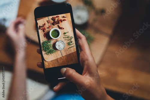 Fototapeta Naklejka Na Ścianę i Meble -  Top view of young hipster girl cooking amateur taking photo on smartphone camera of focaccia recipe ingredients on wood plank to share in cocial networks, Cooking at home concept