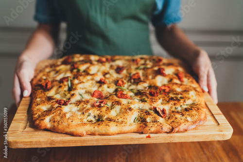 Closeup of woman in green apron holding fresh baked homemade Italian focaccia with rosemary and sun-dried tomatoes on wooden board, Homemade Food Concept