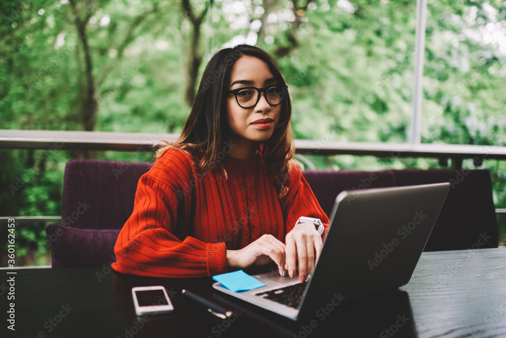 Portrait of young woman in eyeglasses typing on laptop searching information in networks, pensive copywriter looking at camera while updating software and antivirus system on modern netbook Portrait of young woman in eyeglasses typing on laptop searching information in networks, pensive copywriter looking at camera while updating software and antivirus system on modern netbook