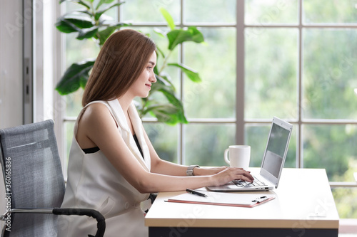 Smiling Asian businesswoman working on a laptop in the office, there is a white cup of coffee on the desk, she felling enjoy, happy working in the workplace