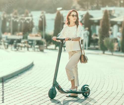 Stylish young woman riding an electric scooter in summer street.
