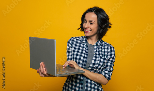 Working day. Half-length photo of a pleased nice girl in a black and white checkered shirt holding her big gray notebook in a right hand and working.