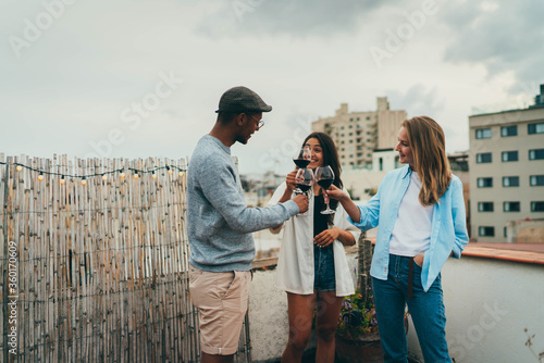 Group of people toasting wine laughing and feeling happy, Friends partying on rooftop enjoying drinks, Outdoor image of Three young friends toasting drinks during rooftop party