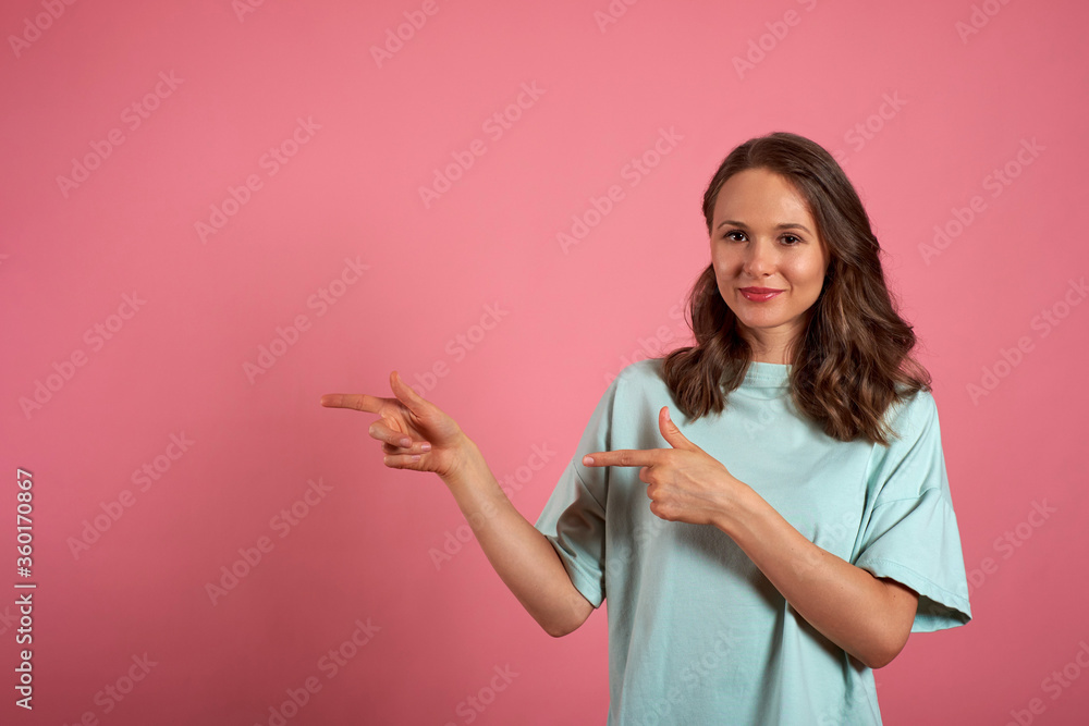 Young woman looks at the camera and pointing somewhere to the side on a pink background Young woman looks at the camera and pointing somewhere to the side on a pink background