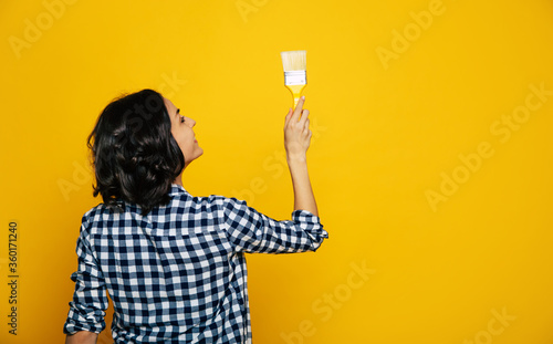 Painting my wall. Close-up length photo of a nice girl, in a black and white checkered shirt, staying back turned, holding a brush in her rigt hand, painting her wall. photo