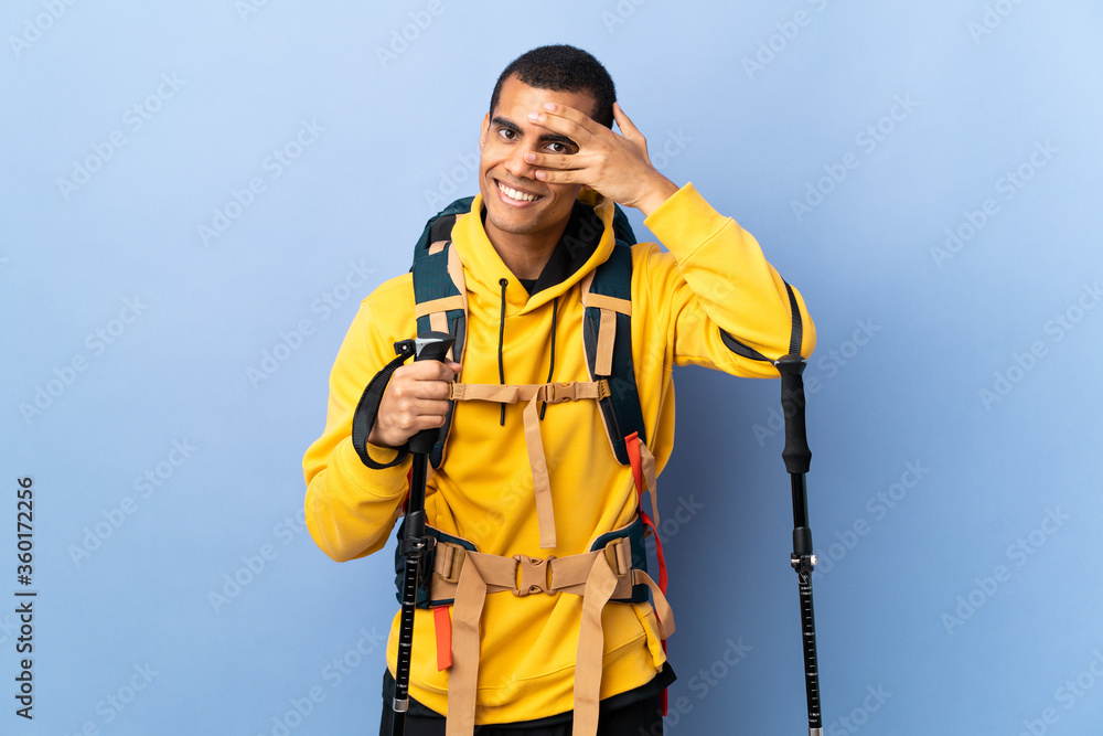 African American man with backpack and trekking poles over isolated background covering eyes by hands and smiling African American man with backpack and trekking poles over isolated background covering eyes by hands and smiling