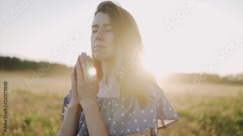 A caucasian beautiful wonan standing on the beautiful field during summer sunset and pray and look at the sky. The woman wears beautiful dress. Close up. SLOW MOTION