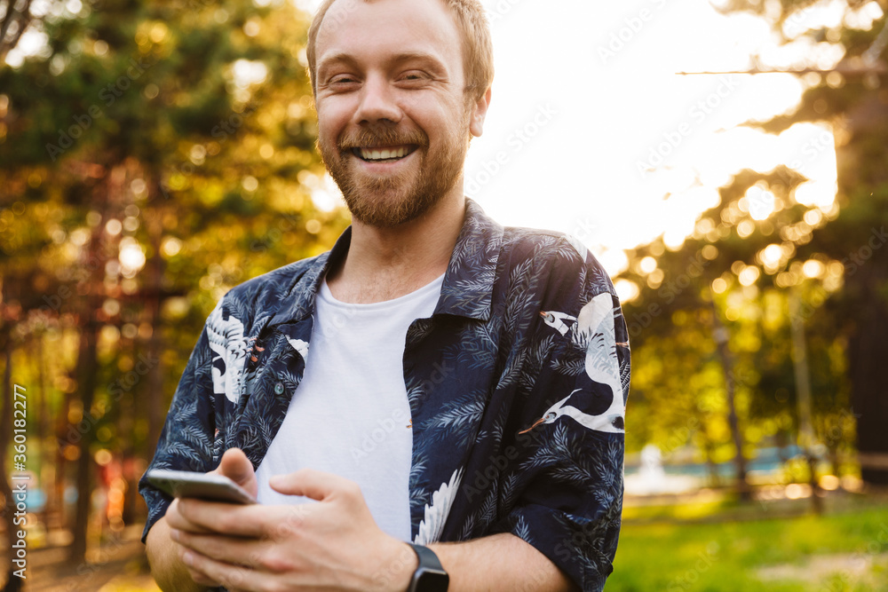 Image of young man smiling and holding cellphone while walking in park Image of young man smiling and holding cellphone while walking in park