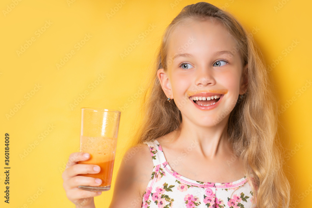 Smiling little girl in summer clothes looking to the side, holding a glass of fresh carrot juice in her hand. Waist shot isolated on yellow, copy space. mustache from carrot juice Smiling little girl in summer clothes looking to the side, holding a glass of fresh carrot juice in her hand. Waist shot isolated on yellow, copy space. mustache from carrot juice