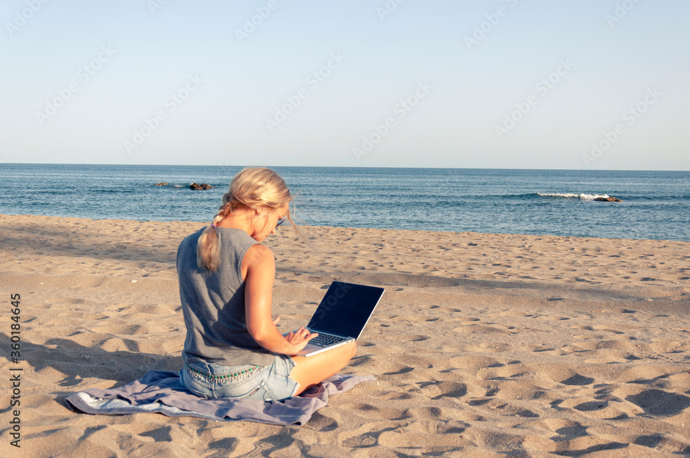 A girl with blonde hair is sitting on a sandy beach against the sea in shorts and a t-shirt and with a laptop, side view