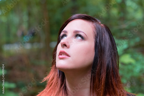 Thoughtful young woman with gray eyes looking up to the sky, wearing nose jewellery, with a stunning hair style in color tones of crimson red and orange, standing in a forest.