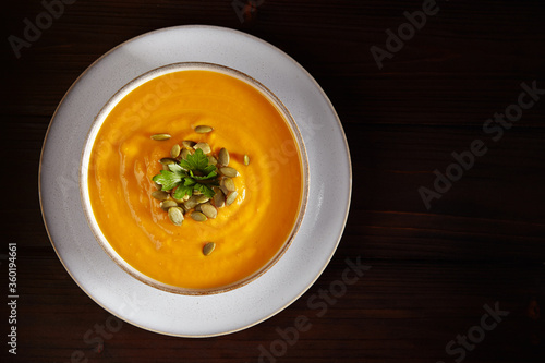 Vegetarian autumn pumpkin soup puree with seeds, greenery and oil in white bowl on black background. Top view.