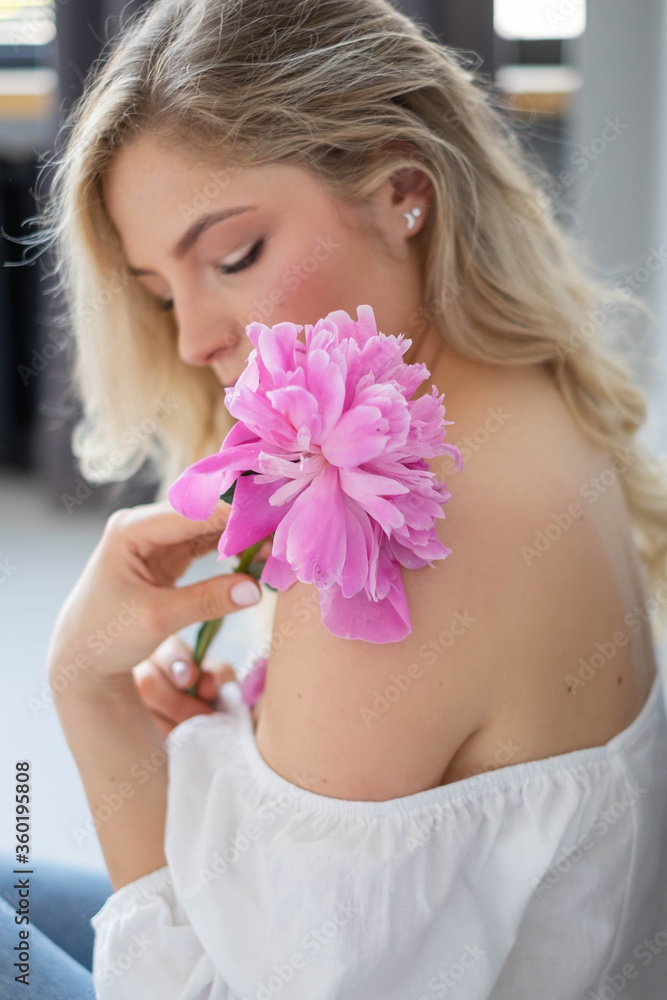 beautiful sensual girl with long blonde hair in blue jeans and a white shirt with a bouquet of pink peonies flowers. Close-up portrait of a woman in studio on a gray background.concept of feminity