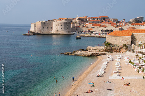 Fototapeta Naklejka Na Ścianę i Meble -  Empty beach club with peach colored sand lapping against the clear waveless azure, turquoise and sea green Adriatic Sea with walled Old Town of Dubrovnik in the background.
