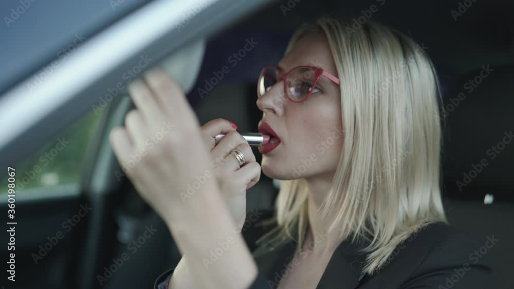 Glamorous blonde in glasses applying the red pomade at mirror in car Glamorous blonde in glasses applying the red pomade at mirror in car